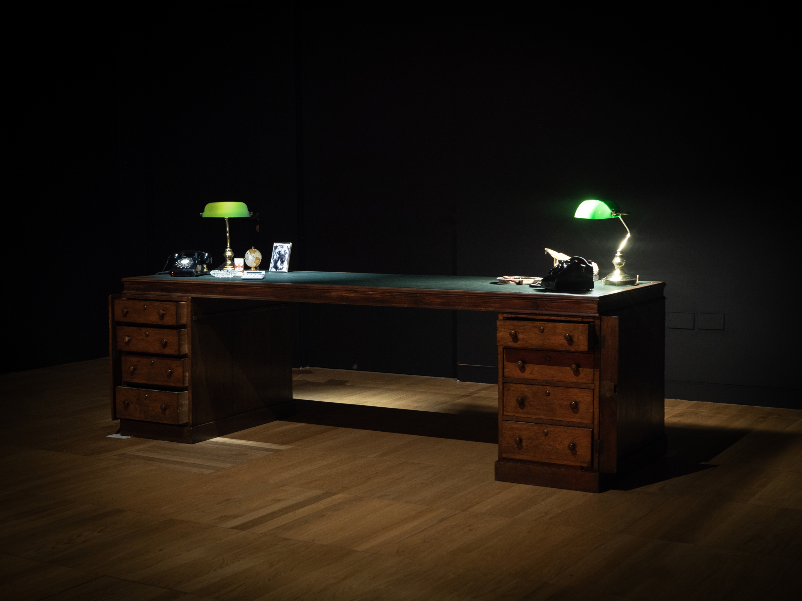 Green, large desk in a dark room with desk lamps at the exhibition Apocalypse Anxieties at Luan Gallery
