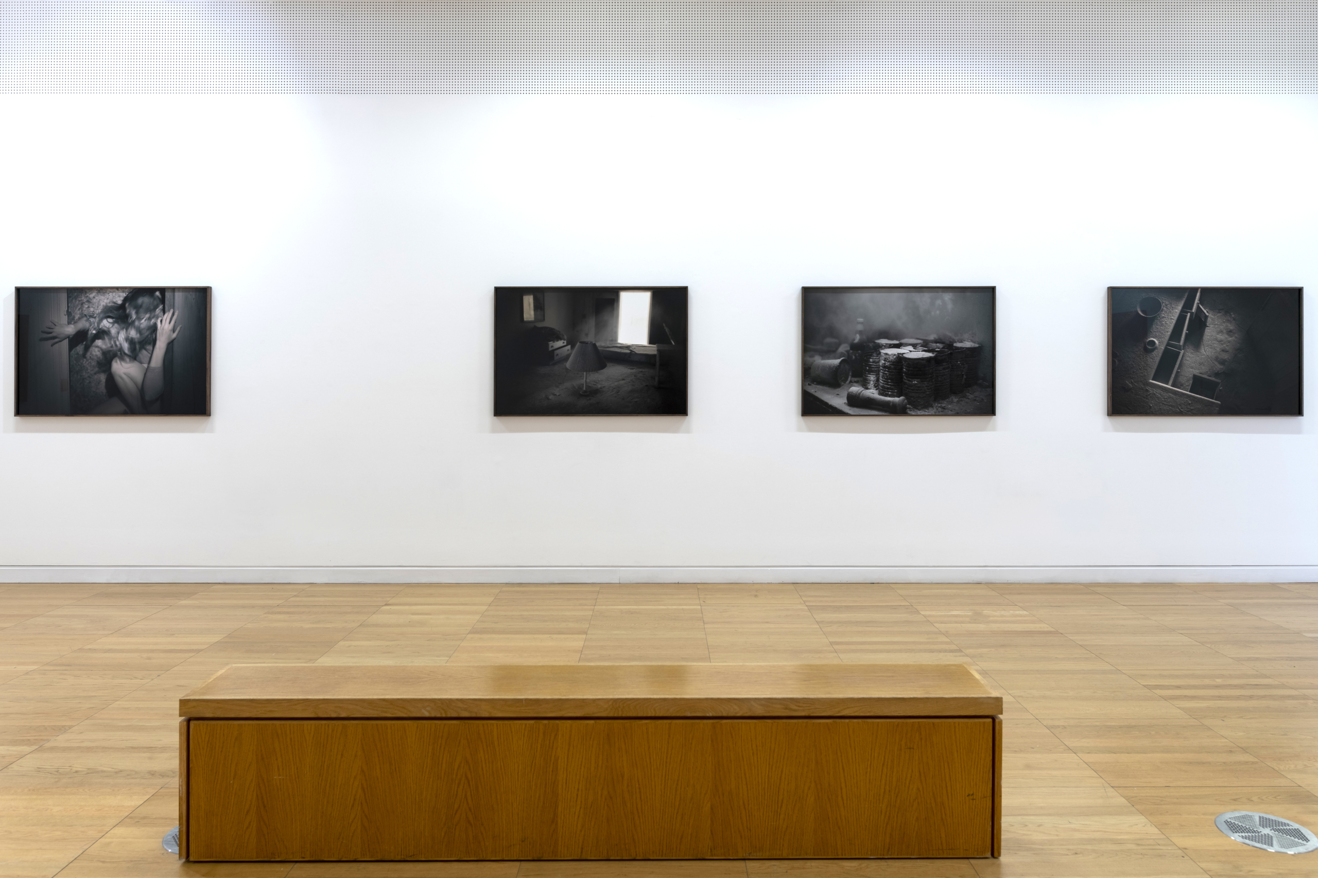 Black and white art on a white wall with a wooden floor and bench at Luan Gallery