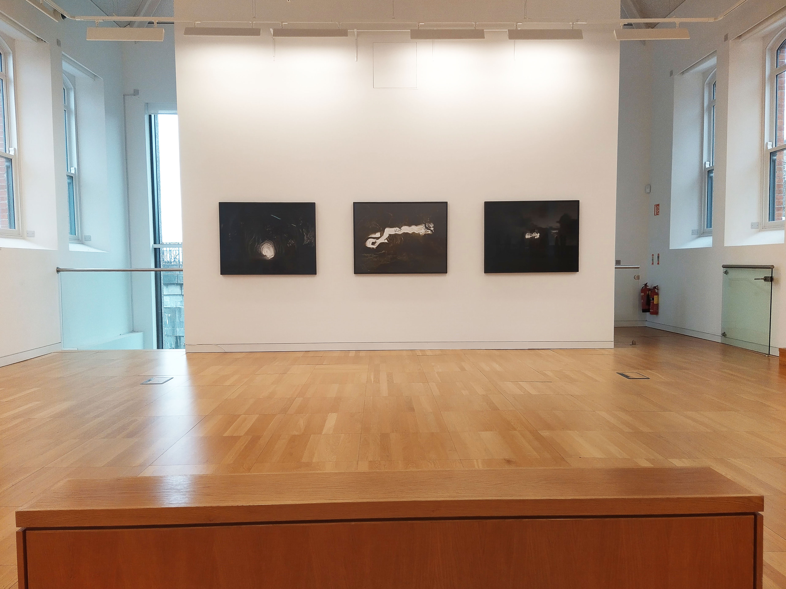 "Nightscapes" exhibition at Luan Gallery - three darker art pieces on a white wall with wooden floor